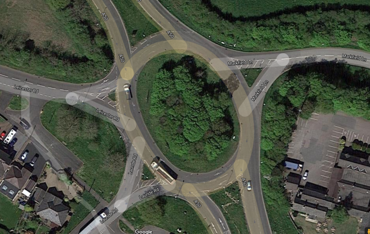 Settle this roundabout dispute - Page 1 - General Gassing - PistonHeads UK - The image shows an aerial view of a complex, multi-lane roundabout intersection. It appears to be a large traffic circle with multiple connecting roads, suggesting it's a significant junction for vehicle movement. The road markings are clearly visible, indicating the designated lanes and directions for drivers. There are also small circles scattered throughout the image, likely representing the locations of various elements at ground level, such as bus stops or street signs. Surrounding the intersection, there is greenery that suggests a mix of urban development and natural areas. The overall scene presents a detailed snapshot of urban planning and infrastructure.