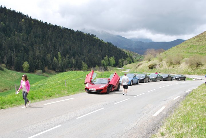 Pistonheads trip to Monaco Historique - Page 2 - Aston Martin - PistonHeads - This image captures a vibrant scene on a curving road set against the backdrop of mountains. On the right side of the road, various cars are parked, possibly on the side of the street. A red car is prominently parked and has its hood up, potentially indicating an ongoing repair or inspection. On the left side of the street, a woman is walking away from the cars, holding a purple book in her hand. The overall atmosphere of the image suggests a picturesque, outdoor setting.