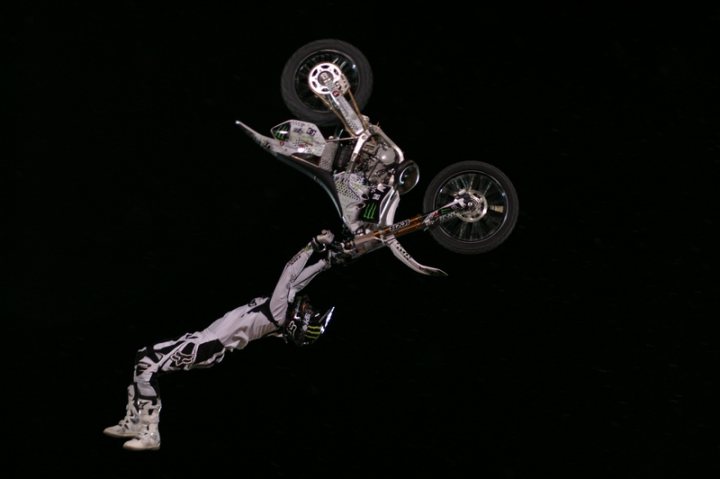 Red Fighters Bull Pistonheads - The image captures a dynamic scene of a person performing a motorcycle stunt to a backdrop of a black sky. The rider is suspended in mid-air, donned in a white motorcycle suit adorned with a black and white pattern, hinting at a possible professional setting. The motorcycle, with its front facing upward, shows two wheels touching the sky, further emphasizing the gravity-defying nature of the stunt. The stark contrast between the rider's monochrome attire and the black sky creates a dramatic visual effect that underscores the thrilling nature of the stunt.