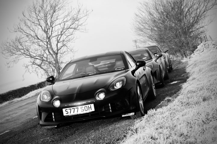 Pistonheads - The image is a black and white photograph featuring two cars parked on the side of a road. There's a noticeable contrast between the lighter foreground and the darker background, creating a sense of depth. Both vehicles appear to be sports cars, with one having a visible license plate that reads "760 SOPHIA". The background reveals trees and a sky, suggesting a rural or semi-rural setting.