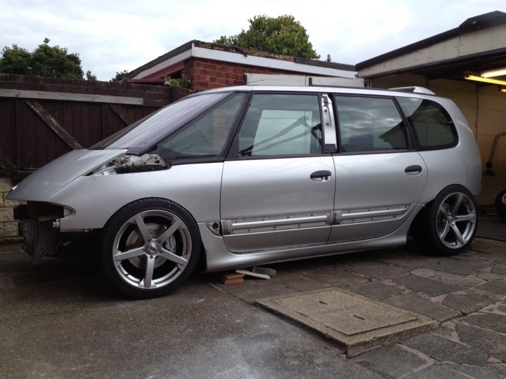 Lexus V8 with NOS in a Renault Espace - yeah lets do it !  - Page 12 - Readers' Cars - PistonHeads - The image shows a silver hatchback parked in front of a garage. It has a unique and visually interesting design, possibly a custom build, with a hood popped open revealing the engine. The hatchback sits on multi-spoke silver wheels, and the body of the car appears to be in good condition with a shiny finish. The background suggests a residential setting with a brick fence and a tree visible.