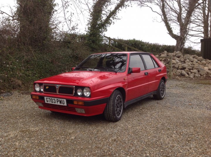 buying an integrale - Page 2 - Alfa Romeo, Fiat & Lancia - PistonHeads - The image shows a bright red car parked on a gravel surface with a natural background. The car appears to be a hatchback, characterized by a small rear window and a distinctive side-vent design. The setting includes a mix of grass and bare land, with some trees visible in the background, suggesting a rural or semi-rural location. There's no other vehicle or person visible in the vicinity, implying it might be a quiet area at the time of the photo. The car seems to be parked dynamically, as if it has been moved there by someone or automatically via a system like a self-parking garage or a Driveaway system integrated into the vehicle.