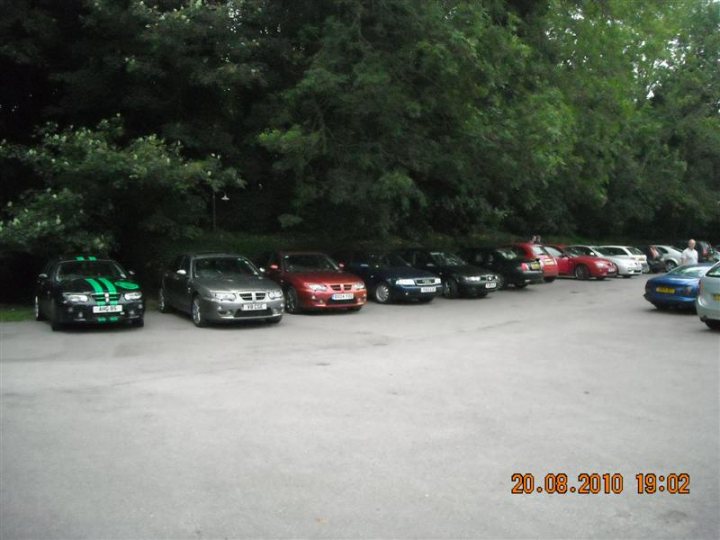 Pistonheads - The image shows a parking lot at night, illuminated by artificial street lights. There is a parking space divider in the foreground, and the cars are parked in various orientations. There are no visible texts, brands, or identifiable features in the image. The photo appears to be taken casually, with a slight tilt to the picture. The atmosphere of the scene is calm and quiet.