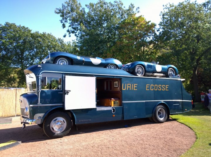 A large truck parked in a parking lot - Pistonheads - The image features a van with a large blue car on its roof. The van itself is blue and black, and it appears to be parked on a gravel or dirt surface. On the side of the van, the word "ECOTEX" is written, suggesting that the vehicle is either part of a company with the same name or is used for eco-friendly purposes. There are also several smaller cars displayed in the back of the van. A bicycle can be seen through the van's rear window, and the van has large black tires with silver hubcaps. The van is parked near a green lawn with a few people visible in the far background.