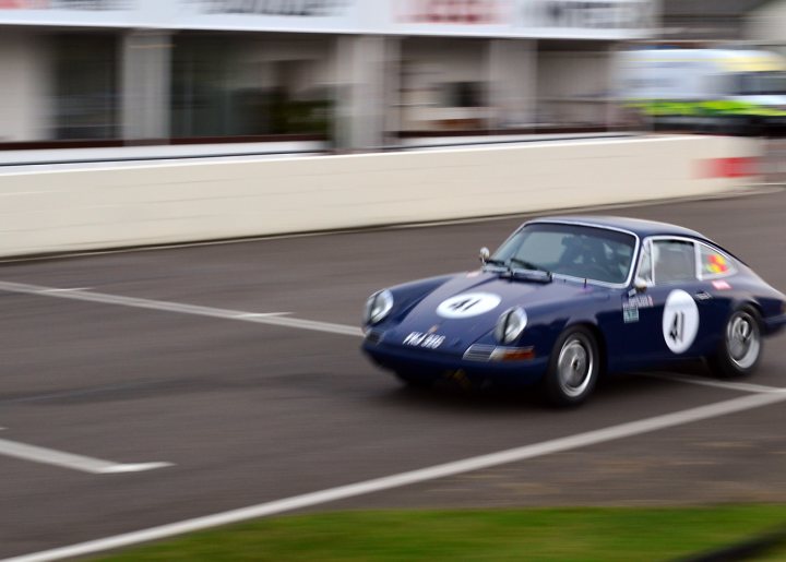 Robert Barrie Track day 24th Feb - photos - Page 1 - Goodwood Events - PistonHeads - The image captures a dynamic scene on a race track. At the center of the frame is a blue racing car, sharply angled towards the left, indicating a high-speed corner. The vehicle carries a white number "11" on its side and a white number "4" on the back, suggesting participation in a racing event. The background reveals a blurred figure of a person and a distant building, adding a sense of depth to the scene. The focus is clearly on the racing car, accentuating its speed and movement.