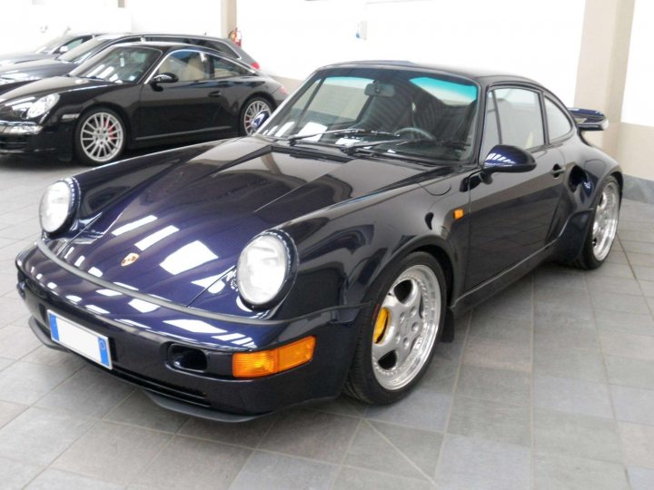 Porsche 964 turbo s lwt - J870 BJN - Page 1 - Porsche Classics - PistonHeads - The image shows a pristine black Porsche parked indoors, bathed in the soft glow of artificial lighting. The car's sleek design is accentuated by a polished surface, gleaming under the reflective tiles of the ceiling. The car's alloy wheels and side mirrors are immaculately clean, reflecting the light in their silvery hue. Another equally elegant car, possibly another Porsche, is visible in the background, adding to the sense of a well-maintained, high-end collection. The indoor setting and the arrangement of the cars suggest a space dedicated to showcasing and preserving these luxury vehicles.