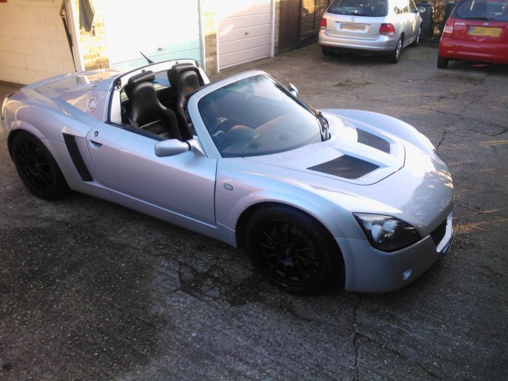 Pistonheads - The image depicts an outdoor scene in a parking lot. The main focus is a sleek, silver, two-door sports car parked on an asphalt surface. The car is quite large, with a design that suggests high performance. Its features include a modern grille, a streamlined hood, and aerodynamic bodywork.

Behind this car are two additional vehicles - a silver car and a red one. The vehicles are parked further away from the camera, partially obscured and not as prominently featured as the sports car in the foreground. The sports car itself takes up most of the frame, highlighting its unique appearance and design. The setting is a parking lot with a clear sky and dry ground visible, indicating it might be a sunny day.