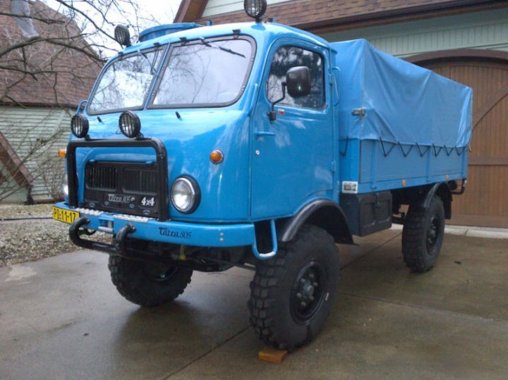 Car model number game - Page 41 - General Gassing - PistonHeads - The image shows a large blue pickup truck parked outside, under a blue tarp. The vehicle is positioned sideways and appears to be of off-road design, as indicated by its high ground clearance and rugged wheels. The front of the truck sports a simple emblem, and the entire vehicle is framed by a building and a tree. A notable feature is the custom headlight on the driver's side, which is more prominent than the standard headlight. The overall scene suggests a working or industrial setting.