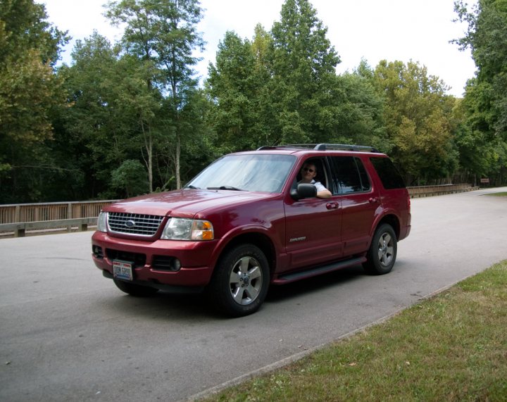 Megamile Pistonheads - In the image, a man is driving a burgundy Ford Explorer on a paved street. The vehicle is presented from a three-quarter front angle, showcasing its front grille, headlights, and the side of the SUV. The man is wearing a white t-shirt and is seen through the open driver's side window, suggesting it's a sunny day. The background is filled with a landscaped area, including a lush green lawn and a line of trees, hinting at a suburban or residential setting.