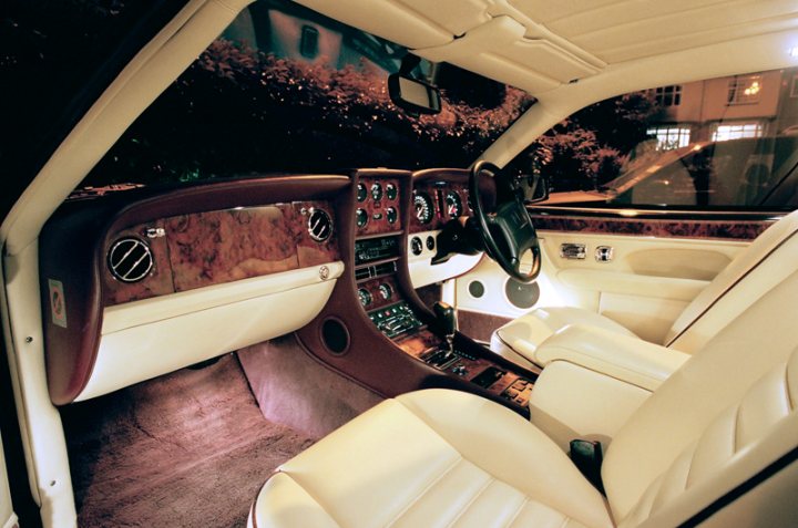 Superb interior lighting - good enough even for photographic - Page 1 - Rolls Royce & Bentley - PistonHeads - The image shows the interior of a high-end vehicle with a harmonious blend of colors and textures. The dashboard is richly adorned with a combination of brown and black leather, punctuated by silver accents and dials. The center console houses a sophisticated sound system, complete with multiple knobs and a hi-fi or speakers, suggesting a focus on audio entertainment. A steering wheel with a similar color scheme is situated on the left side of the dashboard, comfortable leather seats flank each side of the center console. The front passenger seat is vacant, and the atmosphere seems serene and luxurious.