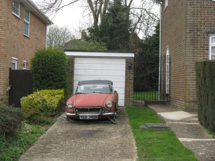 Classics left to die/rotting pics - Page 348 - Classic Cars and Yesterday's Heroes - PistonHeads - This image shows a vibrant, small red car parked on a driveway by the garage of a brick house. The sky is overcast, suggesting an overcast or cloudy day. The yard is neatly trimmed, and there's a well-maintained front lawn. The house appears to be a single-family home with a garage. The overall scene is residential and serene.