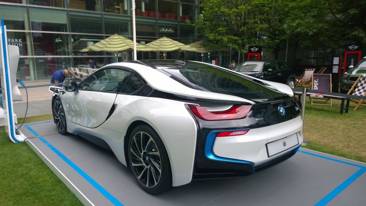 Tesla and i8 at Canary Wharf motorshow this week - Page 1 - EV and Alternative Fuels - PistonHeads - The image features a sleek, white electric car with a distinctive front design. The vehicle's side is adorned with gold accents and the BMW emblem on the trunk. The most eye-catching feature is the vibrant brake light on the back, which transitions between red and blue, adding a touch of technological flair. The car is parked on a gray platform with a blue line marking its spot, which is positioned in front of a store. The setting appears to be an outdoor event or gathering, evidenced by chairs and dining tables in the background.