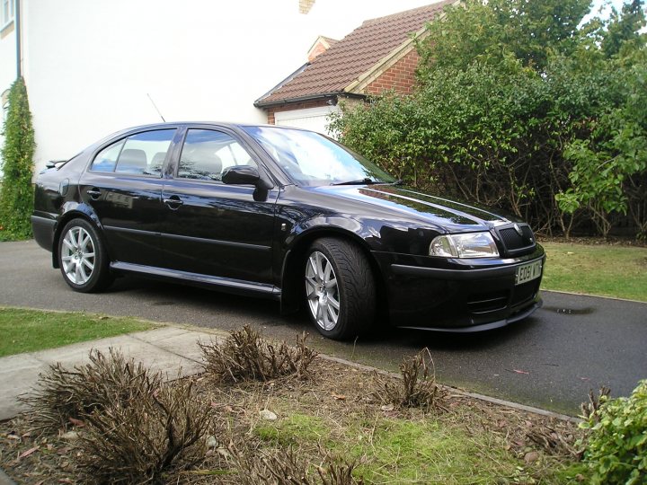 Bit Boost Pistonheads Family - The image captures a tranquil outdoor scene featuring a dark-colored, four-door sedan parked on a wet driveway. The car appears to be a model from the Volkswagen Group, given its distinctive design. Directly in front of the car is a well-maintained garden area, and off to the side, there is a side mirror and a white-colored house with a brick exterior. The overall atmosphere of the image is serene and orderly.