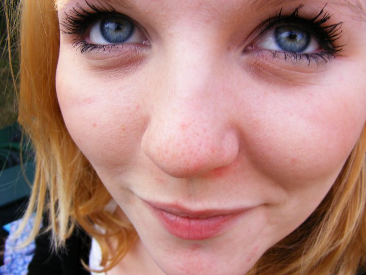 Random Pistonheads Portraits - The image features a young woman with striking blue eyes and long red hair. She appears to be captured in a moment of casual self-expression. Her open mouth suggests she is in the middle of a conversation or possibly smiling. Her blue eyes are the focal point of the image, framed by long eyelashes and a natural, fresh-faced makeup look. The background is blurred, putting the focus entirely on her.