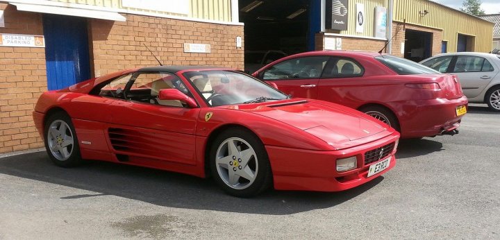 Ferrari 348 chit chat - Page 1 - Ferrari V8 - PistonHeads - The image shows a red Ferrari sports car parked on a paved surface next to a yellow brick building. The car has a sleek design with a black roof, and the setting suggests it might be at a garage or a specialized car workshop. There's another red car visible in the background, but it is not as prominently featured.