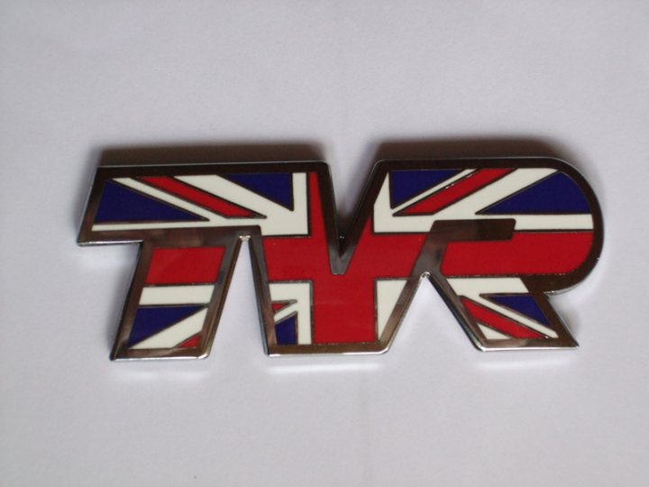 Pistonheads - The image shows a three-dimensional, metallic pin or badge on a white background. It features a prominent letter "T" in a bold, blocky font, which is part of the acronym "TVR." Below the "T," there's a color gradient transitioning from a rich red hue towards a darker area, then to a striking white filled with a graphic of the Union Jack, giving the impression of the British flag (with the cross of Saint Patrick), symbolizing the potential distinction of the pin as related to the United Kingdom or Ireland. The metal appears to be silver or chrome, with a shiny finish. The overall design suggests it could be a lapel pin or an accessory with specific cultural or affiliative significance.