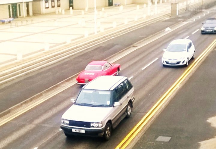 - Page 484 - North West - PistonHeads - The image captures a bustling city street, bathed in the brightness of daylight. Three cars, each distinct in color and model, are the main subjects of this urban scene. A red car, appearing slightly ahead of the other cars, leads the way, followed by a white SUV. A gray car trails behind, maintaining a safe distance. The street is well-marked by double yellow lines on each side, indicating a no-passing zone. The buildings and sidewalks lining the street provide a stark backdrop, typical of urban landscapes.