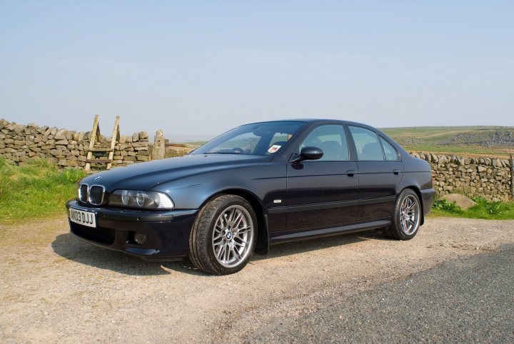 Carbon black or sapphire black - Page 1 - BMW General - PistonHeads - The image depicts a black BMW parked on a dirt path. The car is positioned against the backdrop of a stone wall and a rural landscape, possibly indicating a countryside setting. The sky above is clear and blue, suggesting it might be a calm, sunny day. The foreground of the image captures the details of the BMW, which is captured at an angle that highlights its design and road presence.