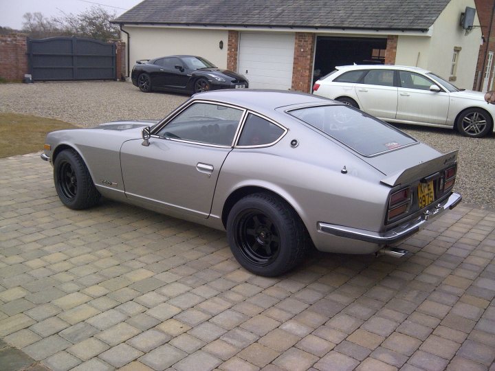 any 240z owners here? - Page 1 - Jap Chat - PistonHeads - The image features a gray sports car parked on a brick driveway in front of a garage. Nearby, additional vehicles are parked, including a blue sports car and a white pickup truck. The garage has a brick exterior, and the area appears to be a residential property. The sky above is overcast, and there are no visible people, focus shifting instead to the vehicles.