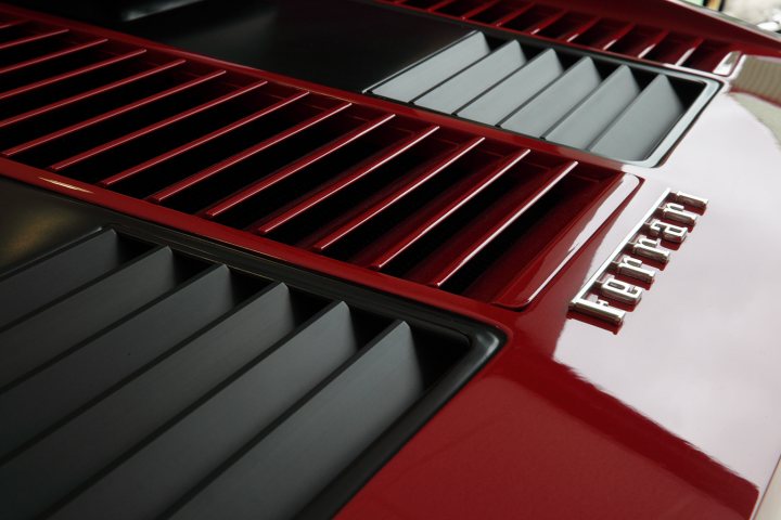 The Berlinetta Boxer thread - Page 1 - Ferrari Classics - PistonHeads - The image shows a close-up view of a large, red ventilation grate. The grating features a series of air holes that are designed to facilitate airflow. The metal has a shiny surface, reflecting the ambient light. In the background, there's a blurred logo with the word "Ferrari" written on it, suggesting that the vent may be part of a Ferrari vehicle. The vent appears to be closed, covering the air passages.