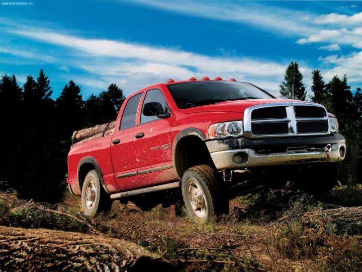 Anyone Driven A Dodge Ram Off Road? - Page 2 - Off Road - PistonHeads - The image features a rugged, off-road style pickup truck perched on a steep hill under a bright blue sky. It is a red Dodge Ram 1500 with a visible bed, equipped with four raised, towing racks and fog lights on the front grille, suggesting readiness for heavy-duty work. The truck is caked in mud, indicating recent use in a difficult terrain. The surrounding area seems to be a dense forest or a disaster area with scattered debris and brush. The terrain's steepness implies rough driving conditions, potentially leading to field tests or risky situations for the vehicle. The overall scene suggests a theme of toughness and ruggedness.