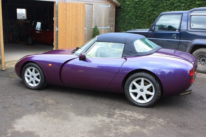 Griff Exterior Colors - Let's See Yours - Page 4 - Griffith - PistonHeads - The image shows a purple convertible parked in a gravel area near a wooden shed. The car has a convertible top down, revealing its two-seat interior. Behind the car, there are other vehicles and a truck, implying a mechanical or storage area. The surrounding greenery suggests an outdoor or semi-rural setting.