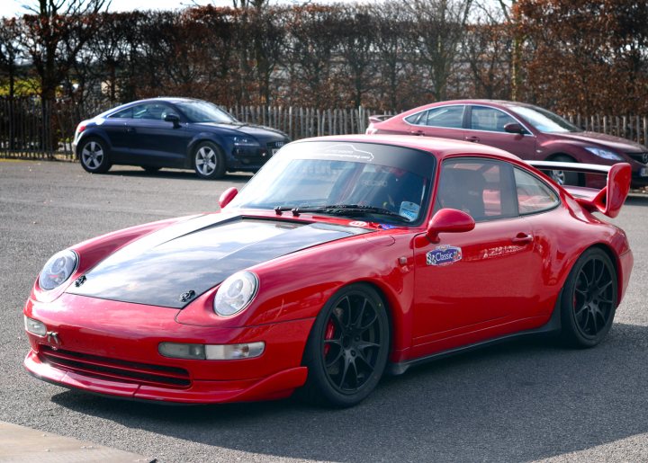 Robert Barrie Track day 24th Feb - photos - Page 1 - Goodwood Events - PistonHeads - In this image, we see a sleek red Porsche parked on the side of a road. The car has a convertible top and a side-exit exhaust, giving it a distinctive and sporty look. There's another car in the background, possibly a blue or black sedan, but it's less noticeable compared to the vibrant red of the Porsche. The setting appears to be an urban or suburban area with trees visible in the background, suggesting it might be a residential or park-like environment.