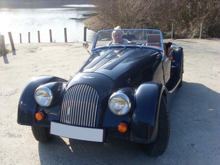 Can I buy clear indicator lenses for this Morgan? - Page 1 - Morgan - PistonHeads - The image features an elderly woman in the driver's seat of a vintage car, which appears to be an expensive antique Bentley. The car is black with a luxurious chrome grill, and the woman seems to be either smiling or squinting in the bright sunlight. The car is parked on a light-colored, visually contrasting gravel lot, possibly in a driveway or off-road area. The setting appears to be a clear day near a body of water, suggesting a peaceful, outdoor environment. The presence of the antique car and the warm weather give the image a nostalgic and leisurely ambiance.