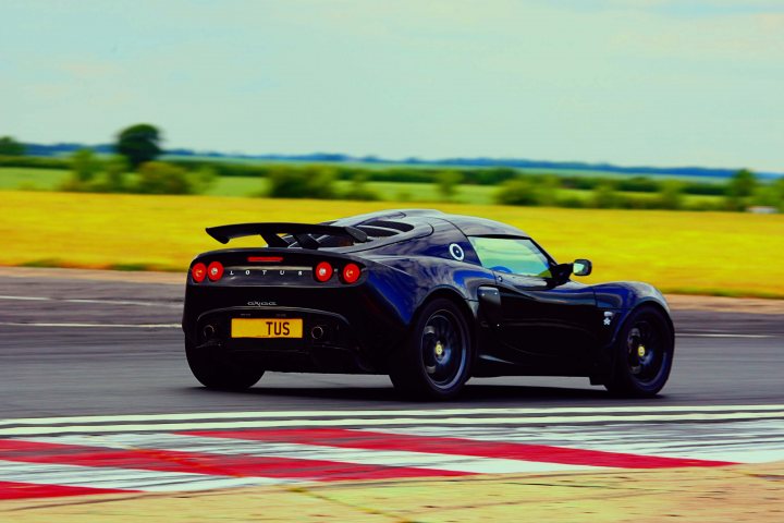 The big Elise/Exige picture thread - Page 6 - Elise/Exige/Europa/340R - PistonHeads - The image depicts a single black sports car speeding down a dark road. This car has a prominent rear wing, adding to its aerodynamic design. The vehicle features a yellow license plate, reading "TUS," which stands out against the black exterior. In the background, there are lines of trees and fields, suggesting an open or rural area, and the sky above is a clear blue with a few clouds.