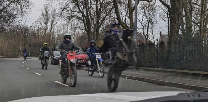 Plague of Electric motorbikes (Sur-Rons) beginning ? - Page 1 - General Gassing - PistonHeads UK - The image captures a lively scene of people riding motorcycles down a road. There are four riders visible, all seemingly in motion, creating a dynamic and energetic atmosphere. They appear to be on a street lined with trees, suggesting an urban setting or perhaps a suburban area. The motorcycles themselves vary in color and style, adding diversity to the scene. The sky above is overcast, casting a soft light over the entire image. In the foreground, there's a car parked by the side of the road, providing a sense of scale and context for the viewer.