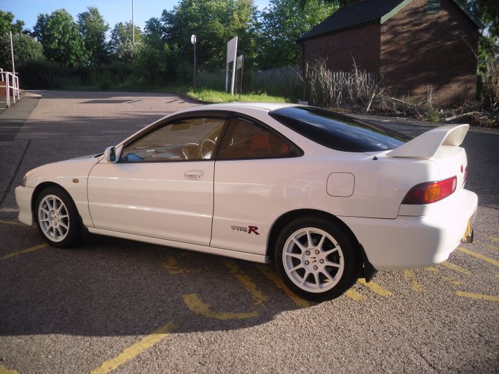 Pistonheads - The image showcases a two-door white sports car, prominently positioned in the foreground. The car features a rear spoiler, adding to its aerodynamic design. It's parked in a car park, which is adorned with yellow paint and signposts, suggesting a controlled parking area. A brick building can be seen in the background, subtly complementing the scene. The sky, visible above, adds a natural element to this otherwise man-made setting.