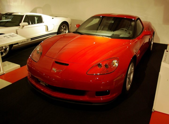 Vettes Nice Pistonheads Place - The image features a vibrant red sports car prominently displayed in what appears to be a showroom or exhibition space, with polished black flooring and a sleek, sculpted design indicative of high-performance vehicles. The car is parked next to a white sports car with a similarly slick design, both side by side on a black-and-white checkered floor. The lighting in the background casts subtle reflections on the cars, adding to the stylish ambiance of the display.