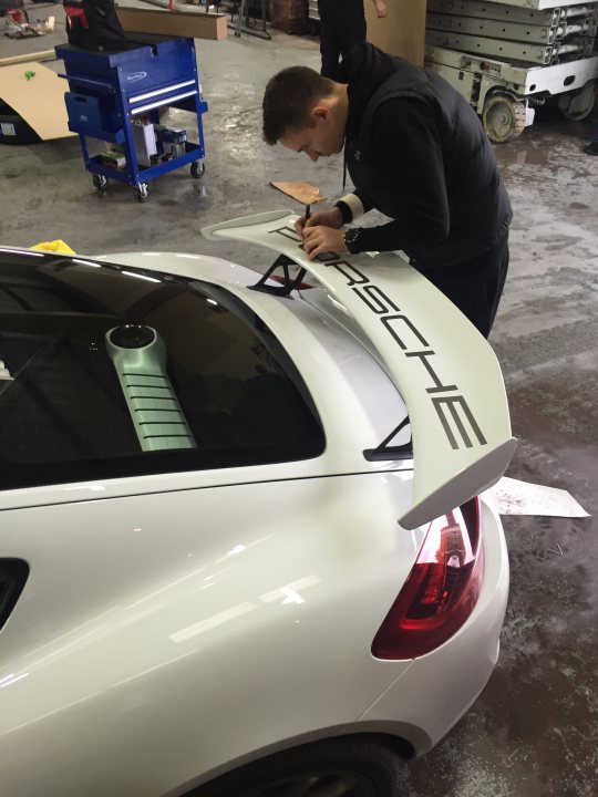 Where to buy these P O R S C H E decals for GT4 wing - Page 1 - Porsche General - PistonHeads - This image captures a garage scene where a man in a black jacket is attentively working on a white Porsche car. His focus is on customizing a large wing or spoiler mounted on the trunk of the car, possibly signing or writing on it. The Porsche logo is prominently displayed, emphasizing the brand of the vehicle. The environment looks like a professional garage or work area, with various pieces of equipment scattered around.