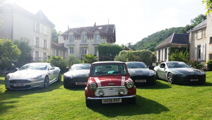 Mini in France - Page 1 - Classic Minis - PistonHeads - This image captures a serene scene in a suburban setting. Six cars, five sports cars and one small car, are parked on a green lawn in front of a residential building. The lawn is well-maintained and adds a touch of nature to the urban landscape. Behind the cars, the building has a large outdoors space, possibly a porch or balcony, and is nestled amidst a backdrop of trees and foliage. The light appears to be from either early morning or late afternoon, giving the setting a warm and inviting atmosphere.