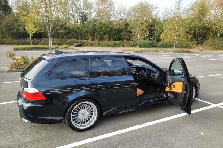 RE: Alpina launches 621hp B5 saloon and estate - Page 3 - General Gassing - PistonHeads - The image depicts a dark blue Audi station wagon parked in a parking lot. It is an outdoor setting with trees and a clear sky visible behind the vehicle. The car has a distinctive black and white wrap on its side, which is partially open, revealing the original color of blue. The car's rear door is swung open, giving a clear view into its interior. There are no people or other vehicles in the immediate vicinity of the Audi.