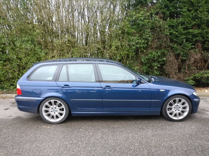 2004 e46 BMW 330i Sport Touring 6 speed - Page 1 - Readers' Cars - PistonHeads - The image shows a dark blue BMW car parked on an asphalt surface. It appears to be a four-door sedan with five-spoke wheels. The vehicle has a dark tint and a manual transmission, as indicated by the gear shift lever visible just inside the driver's window. Behind the car, there is vegetation and a hint of greenery, suggesting the car is parked in an outdoor or suburban setting. The sky is overcast, and there is a reflection of light on the ground, creating a slightly glossy surface where the car sits.