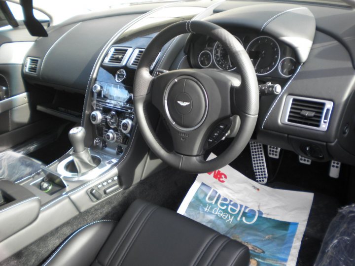 N420 Arrived - Page 1 - Aston Martin - PistonHeads - The image captures the interior of a luxury vehicle, showcasing the driver's side of the dashboard. The driver's seat, partially visible, is plush and black with a contrasting silver center console. Dominating the right side is the steering wheel, a central piece of the dashboard, adorned with a silver emblem. On the left side of the dashboard, the car features a variety of controls and displays, including dials and an infotainment system. In the center console cup holders can be seen. Lastly, a parking tag hangs from the rearview mirror, indicating a recent parking encounter.