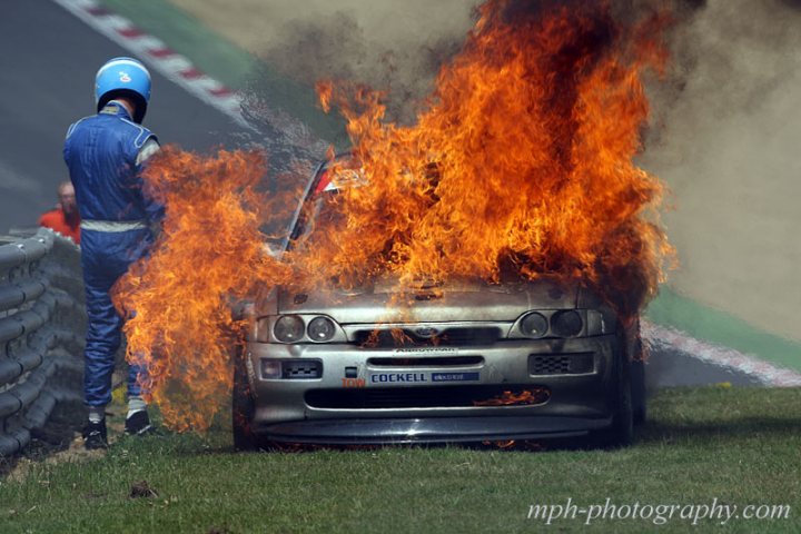 Recommend me a Fire Extinguisher please :) - Page 1 - UK Club Motorsport - PistonHeads - The image captures a dramatic scene at a race track, where a car has caught fire. The flames are so intense that they appear to be rising from the ground, suggesting an explosion from the vehicle. Slowly approaching the fiery wreckage is a firefighter, dressed in a blue uniform and wearing a helmet, presumably to extinguish the flames. The background shows a cityscape with a few trying to rescue. The overall atmosphere is intense and filled with suspense as spectators watch the unfolding disaster.