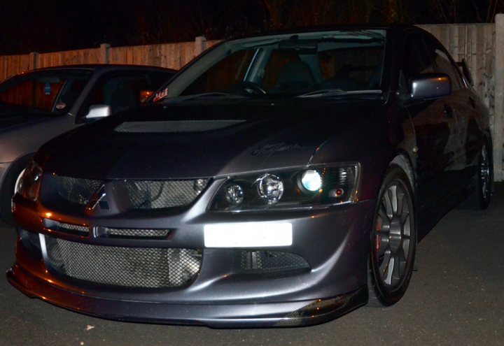 Pistonheads March Essex - This image features a black sports car, which has a glossy finish emphasizing its sleek body. The car is parked outdoors, with a wooden fence visible in the background, and there are other vehicles obscured by the sports car. The car has a prominent front grille and is equipped with aftermarket white round lamps, possibly as replacement headlights, and further modified with a sporty front splitter. The image gives the sense of a personalized and possibly well-maintained sports vehicle.