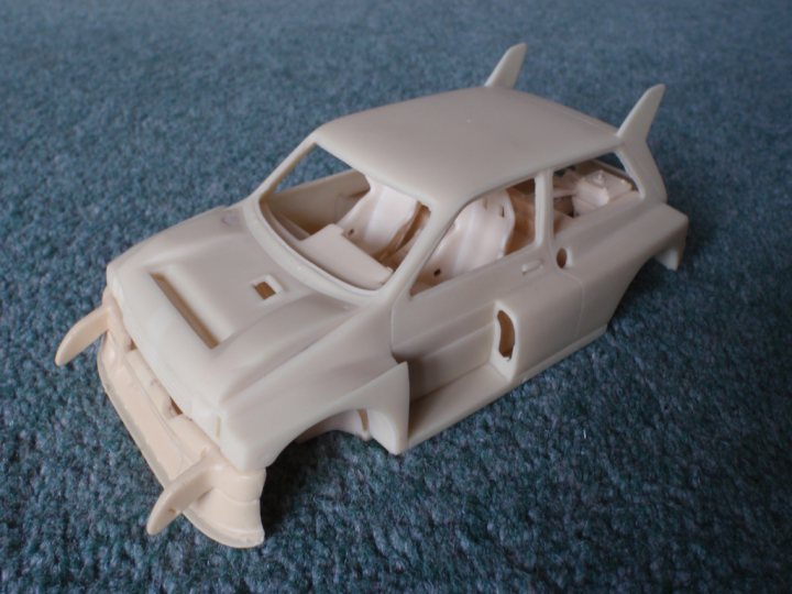 Metro Pistonheads Scale - The image features a white car model laid out on a blue carpeted floor. The car has a distinct design, with raised edges and sloping lines that give it a unique vinyl-like appearance. It's missing its wheels, but the trunk is intact. The attention to detail is evident in the representation of windows and other car parts, suggesting it's a special edition or a custom handcrafted model.