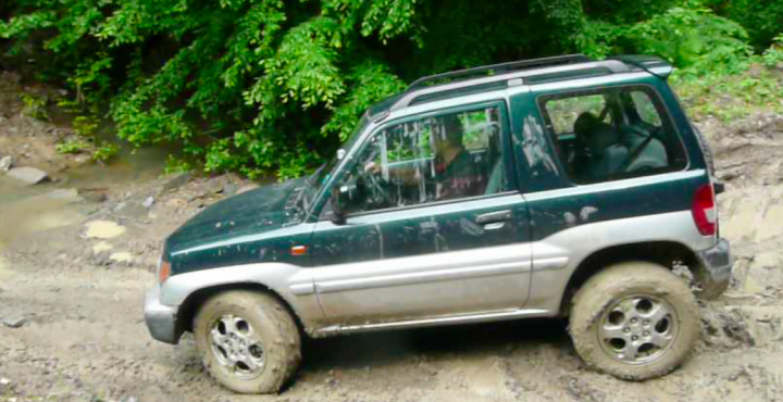 Little overnight trip to the Balkan range - Page 1 - Off Road - PistonHeads - The image features a green sports utility vehicle (SUV) with a silver running board, partially covered in mud, emphasizing a sense of adventure or exploration. The SUV is equipped with an off-road tire and a roof rack, suggesting it's prepared for challenging terrain. The driver appears focused, navigating a muddy path through an environment that looks like a dense forest or jungle. The front view and the mud-filled tires in the foreground further emphasize the SUV's capabilities in such conditions. The image evokes a sense of adventurous travel or a moment of outdoor respite.