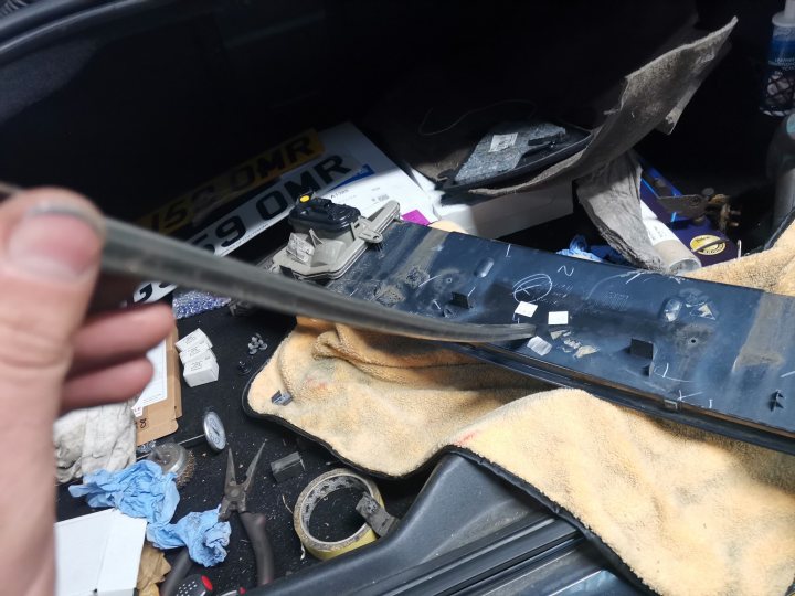 The image shows a close-up of a hand holding an object with a metallic finish, which appears to be a car part. There is a sense of disarray in the background as it looks like parts of a vehicle, including the dashboard and some other items, are scattered about. The focus is on the object being held, suggesting that it might be a tool or component related to repairing or maintaining the car. The setting appears to be indoors, possibly a garage or workshop space.