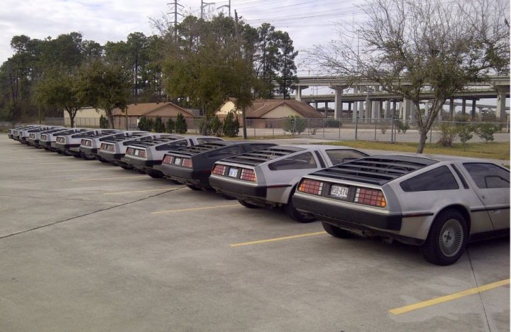 RE: W Motors Lykan Hypercar goes public - Page 2 - General Gassing - PistonHeads - The image shows a row of Delorean cars, an iconic model from the 1980s, parked in a parking lot. The cars are silver with black and orange striping on the sides, and the model looks to be in good condition. In the background, there are trees, a highway overpass, and a lawn with a house across the street. The weather appears to be overcast, with no people visible in the parking lot. The setting suggests a quiet, possibly suburban area, possibly a gathering place for classic car enthusiasts.