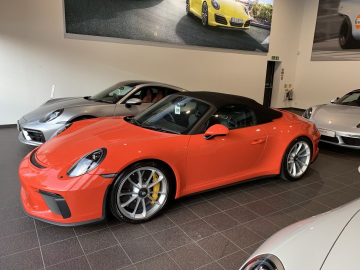 Speedster - Photos and First Impressions  - Page 1 - 911/Carrera GT - PistonHeads - The image shows an indoor setting, possibly a showroom or exhibition, where several sports cars are on display. The most prominent car in the foreground is an orange Porsche convertible with its top down. This vehicle has distinctive features such as alloy wheels and red brake calipers. It's positioned at an angle that allows a clear view of its side profile. In the background, there are additional sports cars, including one in red that appears to be a McLaren. The floor is tiled, and there's a sign with a car image on it, suggesting this is a place where cars are sold or showcased.