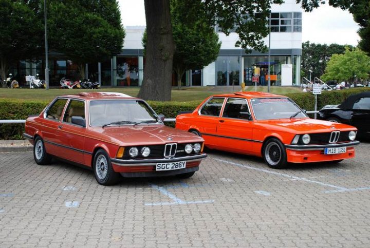 Sunday Pistonheads Service - The image depicts a parking lot with two vintage cars, a red and a white BMW, parked side by side. They feature a classic design with jagged C-pillars and are adorned with Eu license plates, indicating they might be in the European Union. The cars seem to be stationary, parked near a building in the background, under a tree providing shade. The parking lot appears to be busy, as there are several other cars and motorcycles present in the distance.