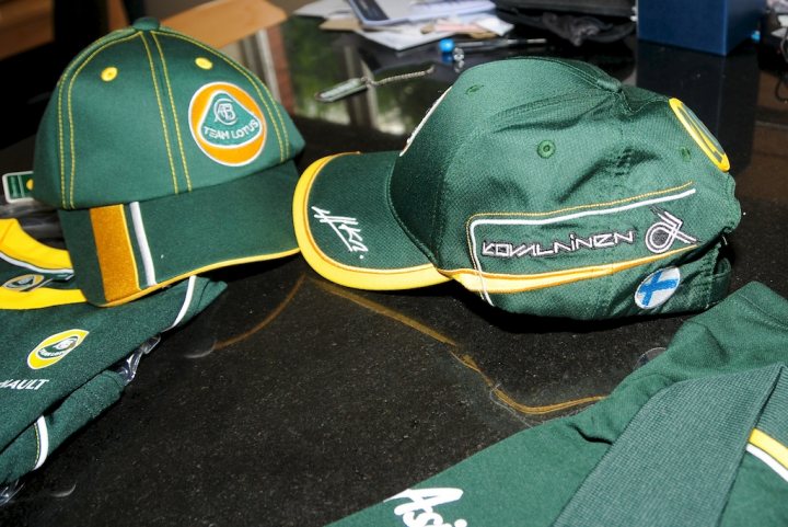 New Team Lotus Gear Turned up today!! - Page 1 - Caterham - PistonHeads - The image shows a pair of green baseball caps placed on a table. They appear to be sports merchandise, with visible logos and text. The one on the left seems to have a circular logo on the front, while the cap on the right has a horizontal stripe with contrasting colors. The area surrounding the table is slightly blurred, suggesting the background is indoors but out of focus. The caps are positioned side by side, with one sitting slightly askew compared to the other.