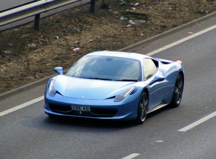 Spotted Forthright MC - Page 36 - Herts, Beds, Bucks & Cambs - PistonHeads - The image shows a vibrant blue Ferrari E4D sports car traveling down a multi-lane highway. The car is positioned slightly to the right on the road, with its front visible as it occupies one lane. The car's sleek design and distinctive headlights are prominently featured. The highway is bathed in daylight, and the shoulder adjacent to the car is bordered by a dusty embankment and scattered with discarded litter.