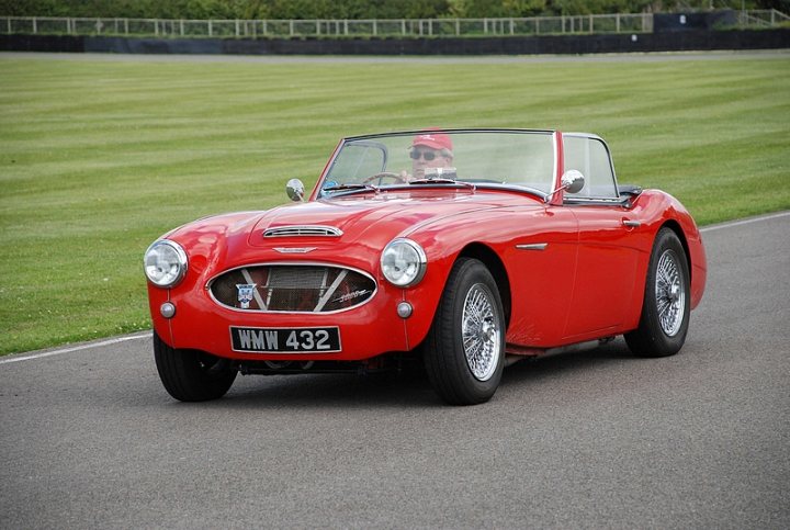 Goodwood Breakfast Club, Pre 1966 classic sunday - Page 1 - Goodwood Events - PistonHeads - The image showcases a red vintage car, possibly a MG, parked on a grassy expanse. It's a sunny day, and there are other cars visible in the background. The perspective of the photo is from the side of the road, with the car's driver's seat visible, suggesting the driver's side door is open. The car's classic design and the person sitting in the driver's seat add a sense of nostalgia and enjoyment to the scene.