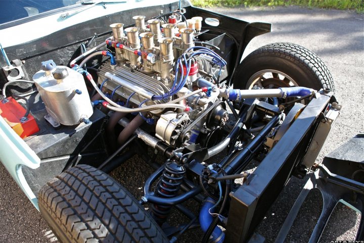 Early TVR Pictures - Page 17 - Classics - PistonHeads - The image showcases a car with an under-car engine visible. The engine has numerous silver components and blue tubes sticking out, suggesting a custom or powerful setup. The car is facing towards the camera, and you can see parts of the hood and a side-view mirror. There are orange cones behind the car, indicating a potential work zone. The ground beneath the car appears to be asphalt. The overall setting suggests a situation where the car's engine is being inspected or serviced.