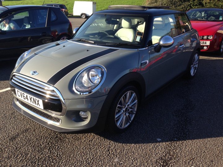 South West Wales Breakfast Meet - Page 143 - South Wales - PistonHeads - The image features a close-up view of a gray Mini Cooper car parked in a lot. The Mini is a small vehicle known for its fuel efficiency and maneuverability, and the car in the image has a distinctive black and silver stripe design on its hood. The side view mirrors are prominently visible, reflecting the surrounding area. In the background, another red car can be seen, parked in the same lot, adding context to a commercial or public parking area. The image captures a moment in time, with the focus primarily on the Mini Cooper car and its striking design feature.